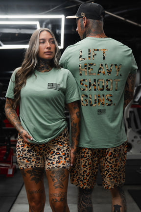 Two tattooed individuals in a gym sport matching Loaded Leopard LHSG - Seafoam Green Tees from Tango Charlie Apparel and leopard print shorts; the bold back of one shirt declares LIFT HEAVY. SHOOT GUNS., proudly expressing their Second Amendment values and commitment to high-performance fitness gear.