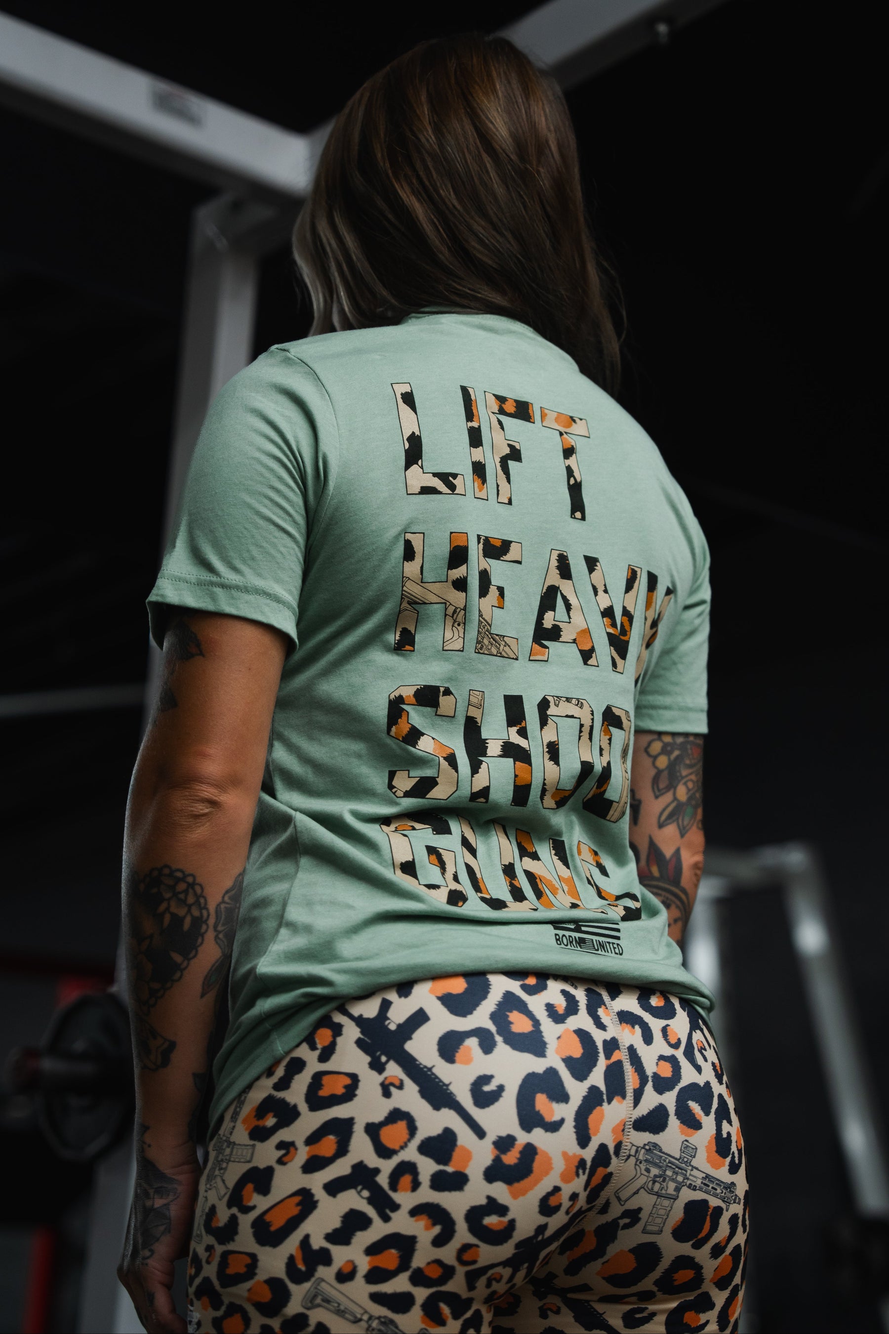 Wearing the Tango Charlie Apparel Loaded Leopard LHSG - Seafoam Green Tee, which proudly features “LIFT HEAVY SHOOT GUNS” on the back, a person stands in the gym with bold leopard print leggings and intense fitness gear, showcasing their support for the Second Amendment.