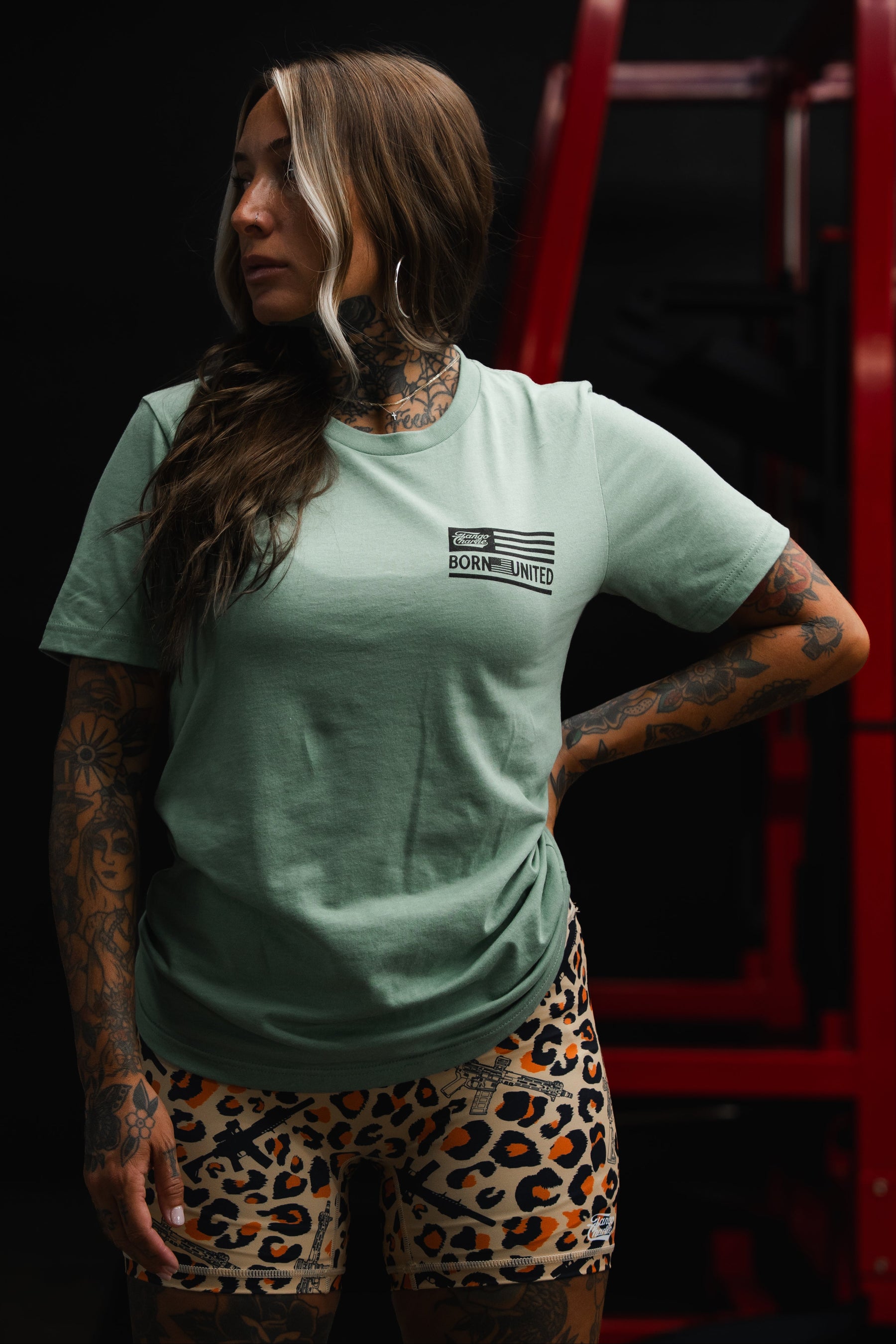 A woman with tattoos stands in front of red gym equipment, looking to the side, wearing leopard print shorts and a Tango Charlie Apparel Loaded Leopard LHSG - Seafoam Green Tee—ideal for fans of Lift Heavy Shoot Guns.