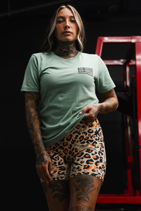 A tattooed woman stands indoors, holding her Loaded Leopard LHSG - Seafoam Green Tee by Tango Charlie Apparel, paired with leopard print shorts. Red gym equipment in the background highlights her high-intensity fitness gear.