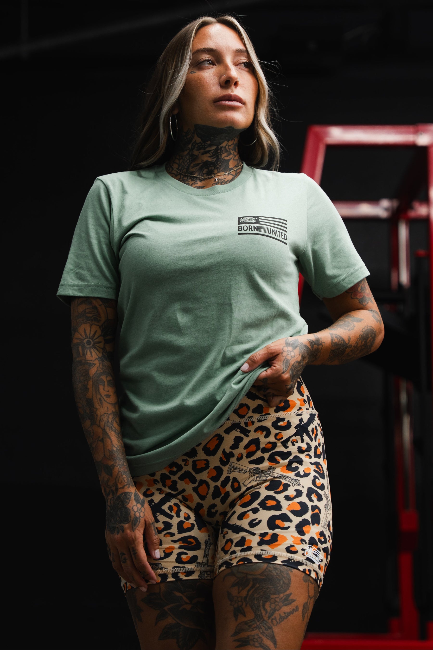 A woman with tattoos stands indoors, confidently holding her shirt while wearing the Tango Charlie Apparel Loaded Leopard LHSG - Seafoam Green Tee and leopard print shorts—a bold look that pairs perfectly with high-intensity fitness gear.