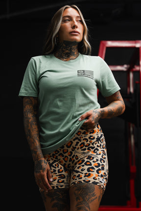 A woman with tattoos stands indoors, confidently holding her shirt while wearing the Tango Charlie Apparel Loaded Leopard LHSG - Seafoam Green Tee and leopard print shorts—a bold look that pairs perfectly with high-intensity fitness gear.