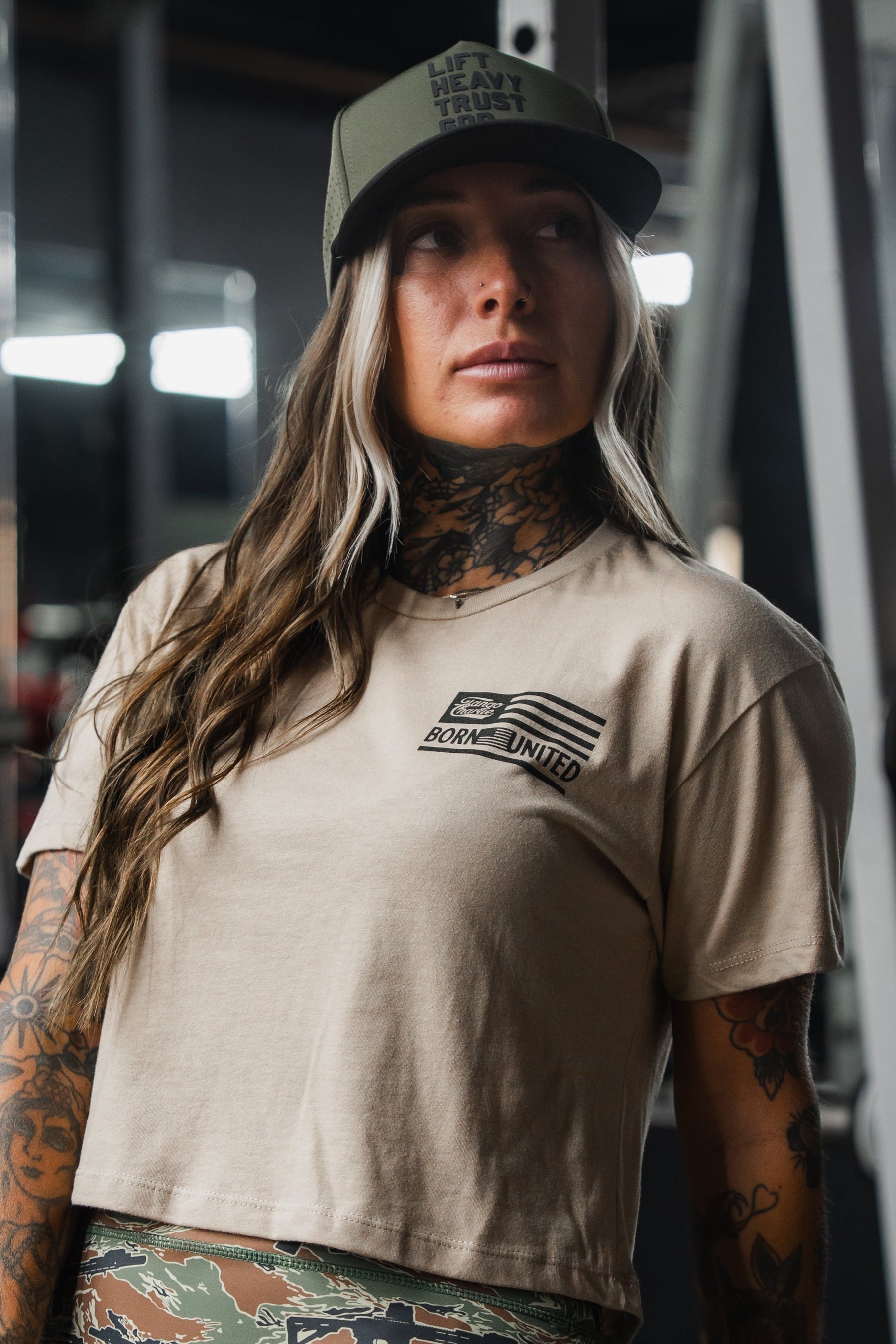A person with long hair and tattoos, wearing a cropped t-shirt and the Tango Charlie Apparel Lift Heavy. Trust God. Hydro - OD Green Snapback Hat, stands indoors looking slightly to the side.