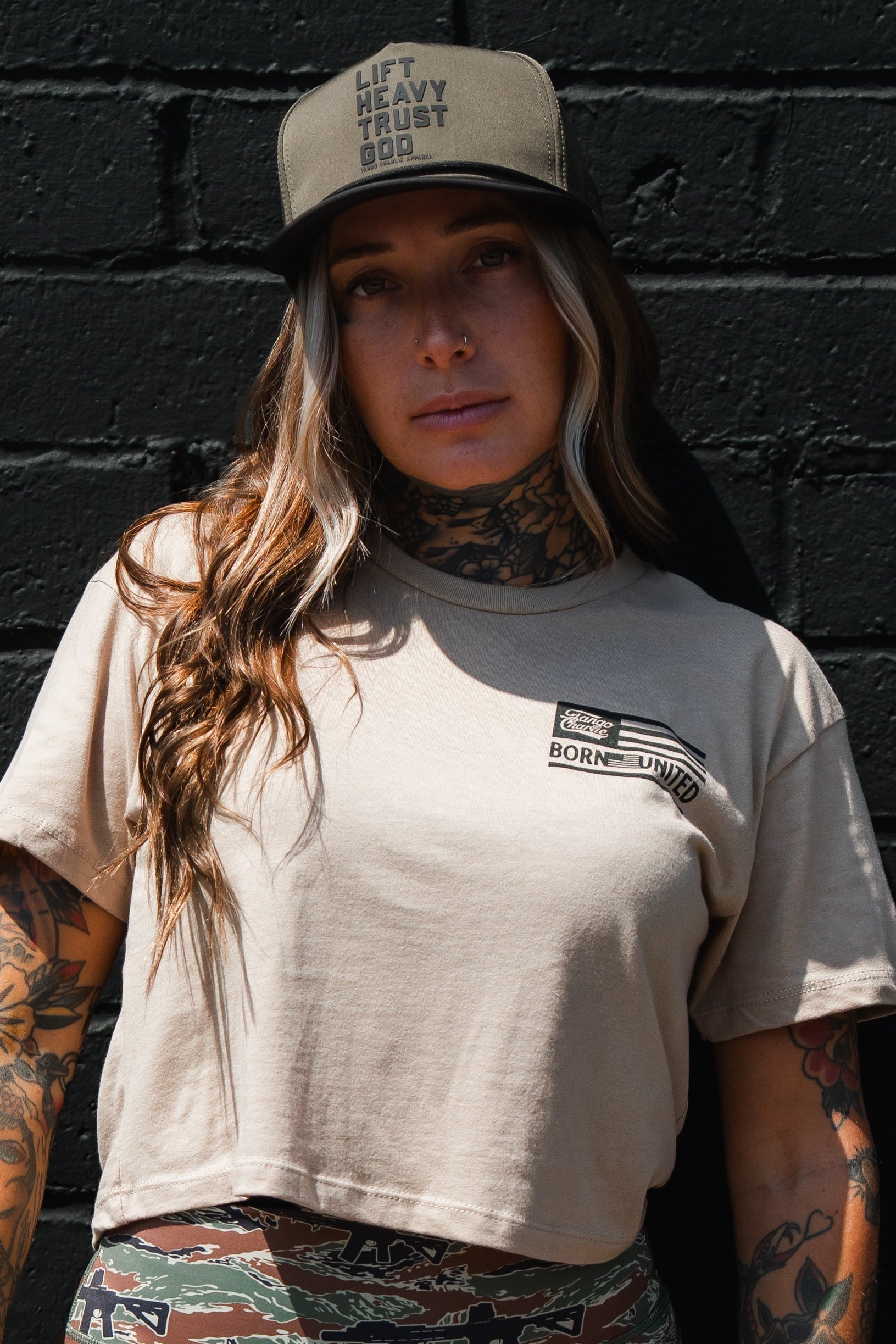 A woman with tattoos wears a beige crop top and a Tango Charlie Apparel Lift Heavy. Trust God. Hydro - OD Green Snapback Hat, standing against a black brick wall.