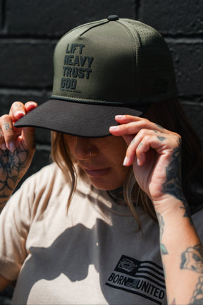 A tattooed individual adjusts an OD Green Lift Heavy. Trust God. Hydro Snapback Hat by Tango Charlie Apparel while wearing a beige Born United t-shirt, standing against a dark brick wall.