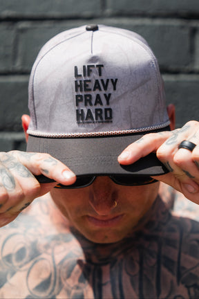 Wearing the Tango Charlie Apparel Lift Heavy. Pray Hard. Deadwood Camo Hydro - Snapback Hat, boldly emblazoned with its inspiring message across the front, a person grips the brim and looks down, showcasing their tattooed arms and nose ring.