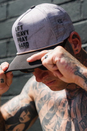 A tattooed individual adjusts a Tango Charlie Apparel Lift Heavy. Pray Hard. Deadwood Camo Hydro Snapback Hat, standing against a brick wall and partially shading their face with the cap.