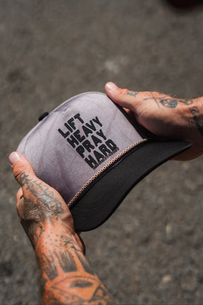 Tattooed hands hold a Lift Heavy. Pray Hard. Deadwood Camo Hydro – Snapback Hat by Tango Charlie Apparel, featuring the words LIFT HEAVY PRAY HARD on the front. This water-resistant hat rests against a blurred, gravelly background.