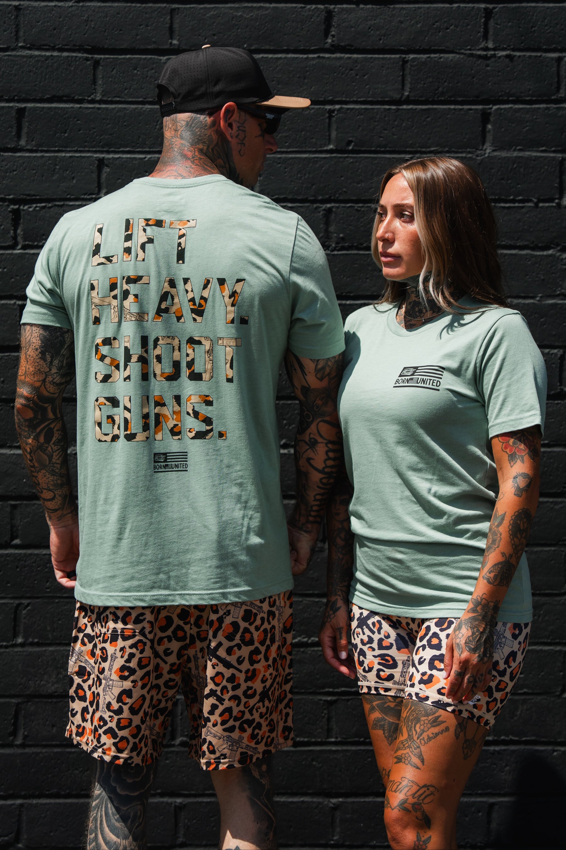 Two tattooed individuals sporting matching Loaded Leopard LHSG - Seafoam Green Tees from Tango Charlie Apparel and leopard print shorts stand before a black brick wall. Embracing their love for high-intensity fitness gear, one shirt features the bold slogan "LIFT HEAVY SHOOT GUNS," reflecting both strength training and Second Amendment pride.