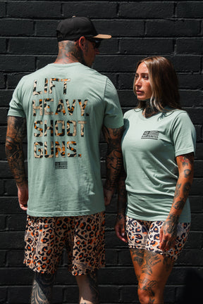 Two tattooed individuals sporting matching Loaded Leopard LHSG - Seafoam Green Tees from Tango Charlie Apparel and leopard print shorts stand before a black brick wall. Embracing their love for high-intensity fitness gear, one shirt features the bold slogan "LIFT HEAVY SHOOT GUNS," reflecting both strength training and Second Amendment pride.