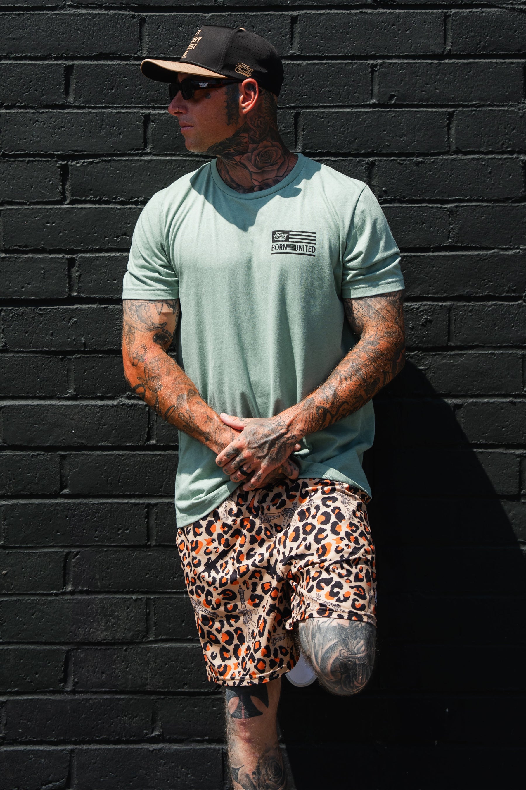 A man with tattoos wearing the Tango Charlie Apparel Loaded Leopard LHSG - Seafoam Green Tee, leopard print shorts, a cap, and sunglasses stands against a black brick wall.