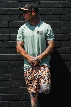 A man with tattoos wearing the Tango Charlie Apparel Loaded Leopard LHSG - Seafoam Green Tee, leopard print shorts, a cap, and sunglasses stands against a black brick wall.