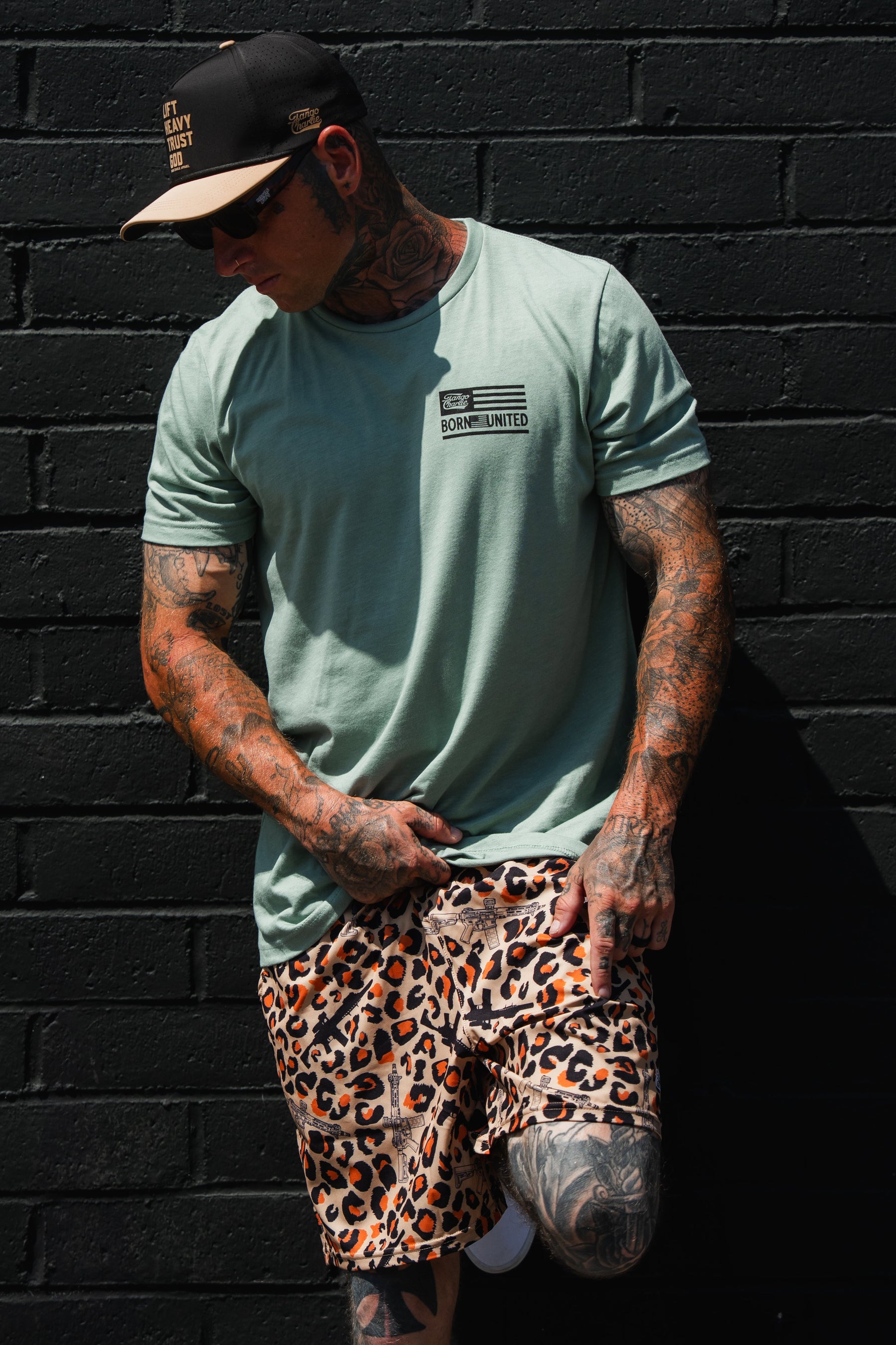 A tattooed individual wearing the Tango Charlie Apparel Loaded Leopard LHSG - Seafoam Green Tee, leopard print shorts, and a black cap stands against a black brick wall, looking down and pointing at their shorts—a bold choice in high-intensity fitness gear inspired by the spirit of Lift Heavy Shoot Guns.