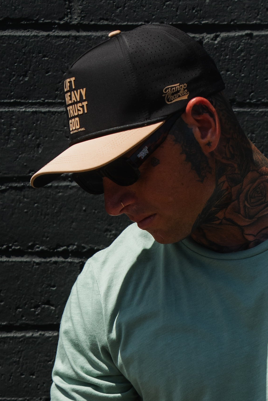 A man in a mint green shirt, sunglasses, and tattoos stands against a black brick wall wearing the Tango Charlie Apparel “Lift Heavy. Trust God. Hydro” Black Snapback Hat.
