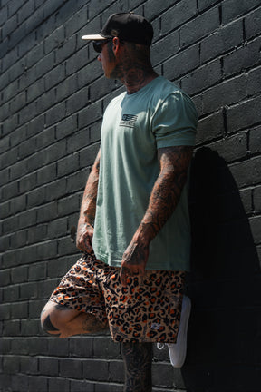A tattooed individual sporting the Loaded Leopard LHSG - Seafoam Green Tee by Tango Charlie Apparel, along with a cap and leopard print shorts, leans against a black brick wall, exuding a bold "Lift Heavy Shoot Guns" vibe with their high-intensity fitness style.
