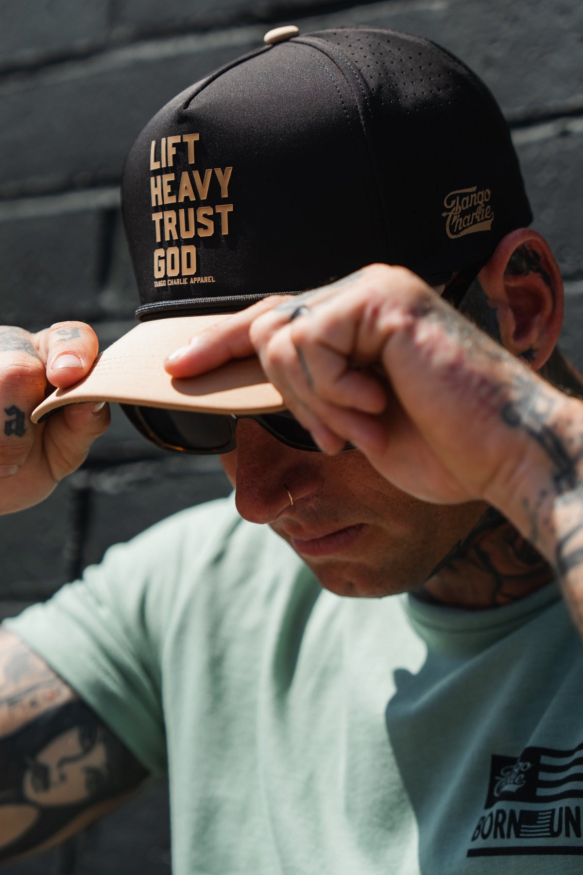 A tattooed individual wearing sunglasses and a light green shirt adjusts the brim of a Tango Charlie Apparel “Lift Heavy. Trust God. Hydro - Black Snapback Hat”—a sweat-resistant cap—while standing in front of a dark brick wall.