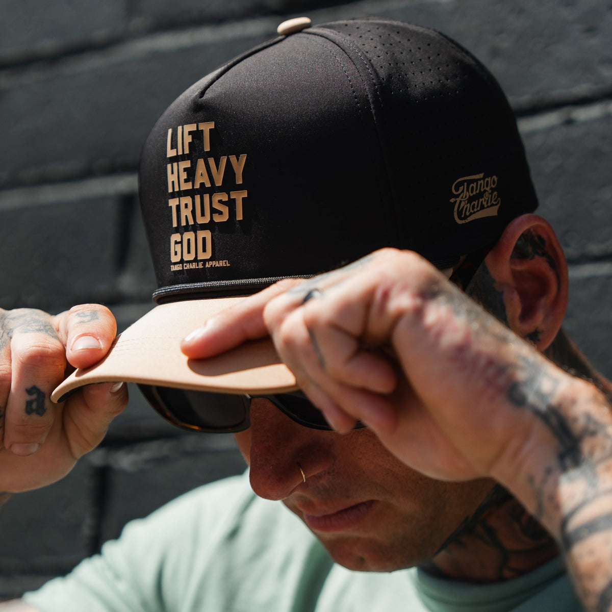 A person wearing sunglasses adjusts a Tango Charlie Apparel Lift Heavy. Trust God. Hydro - Black Snapback Hat, which is designed to be sweat resistant, while standing in sunlight near a brick wall.