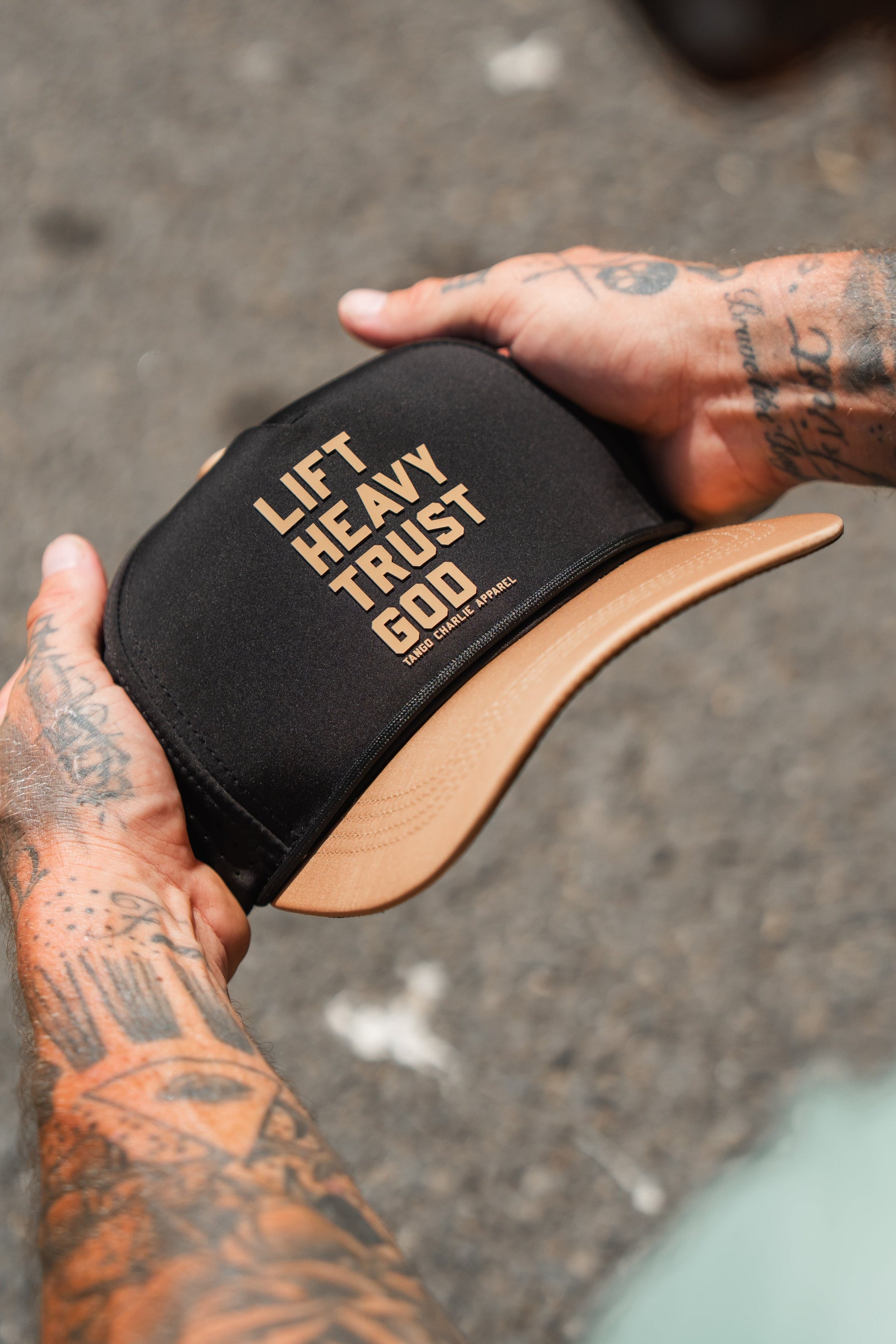 A tattooed individual holds the Lift Heavy. Trust God. Hydro - Black Snapback Hat by Tango Charlie Apparel—a Christian gym hat crafted for both style and purpose, featuring the bold phrase “LIFT HEAVY. TRUST GOD.” across the front. This sweat-resistant snapback is ideal for anyone seeking motivation and faith during their toughest workouts.
