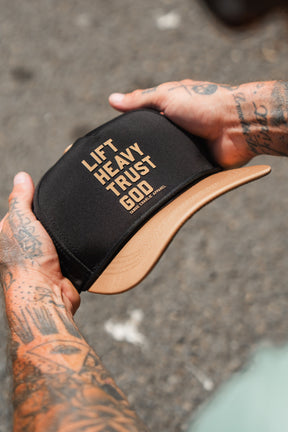 A tattooed individual holds the Lift Heavy. Trust God. Hydro - Black Snapback Hat by Tango Charlie Apparel—a Christian gym hat crafted for both style and purpose, featuring the bold phrase “LIFT HEAVY. TRUST GOD.” across the front. This sweat-resistant snapback is ideal for anyone seeking motivation and faith during their toughest workouts.