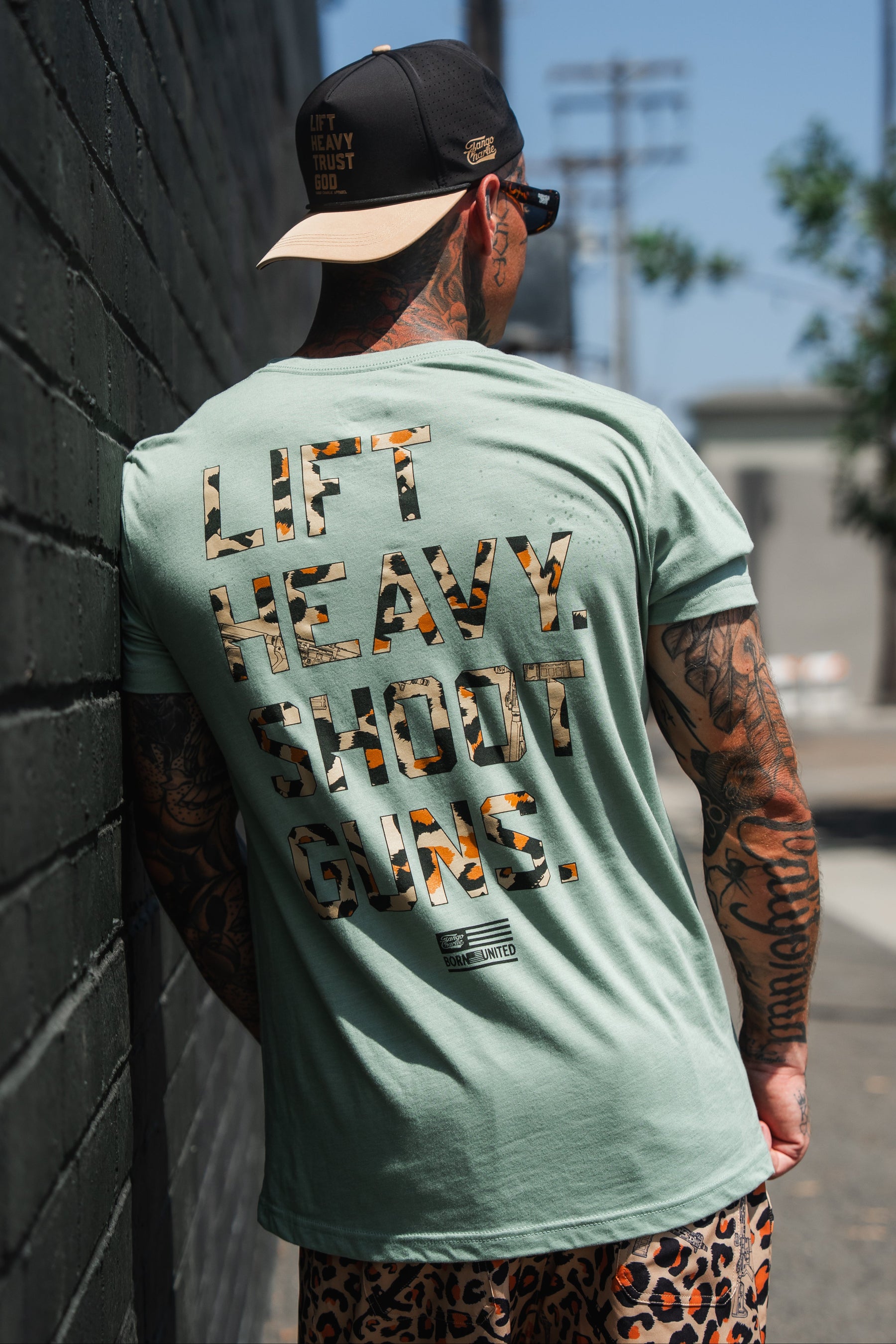 A tattooed man sporting sunglasses and a cap stands by a wall, wearing the Loaded Leopard LHSG - Seafoam Green Tee from Tango Charlie Apparel. The shirt features "LIFT HEAVY. SHOOT GUNS." in large bold letters across the back, making a powerful statement about his dedication to strength and support for the second amendment.