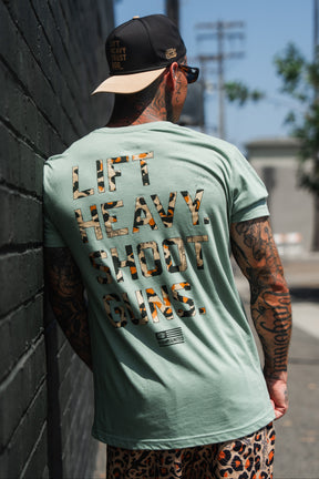A tattooed man sporting sunglasses and a cap stands by a wall, wearing the Loaded Leopard LHSG - Seafoam Green Tee from Tango Charlie Apparel. The shirt features "LIFT HEAVY. SHOOT GUNS." in large bold letters across the back, making a powerful statement about his dedication to strength and support for the second amendment.