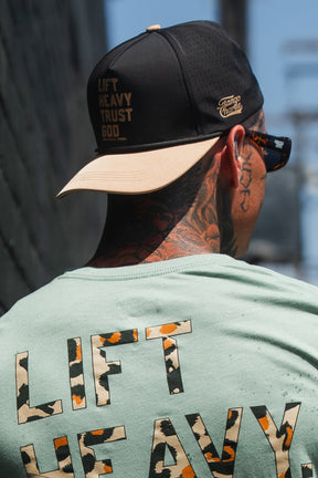 A man with neck tattoos is wearing a backwards black “Lift Heavy. Trust God. Hydro” snapback hat by Tango Charlie Apparel and sunglasses. The sweat-resistant cap features the phrase “LIFT HEAVY TRUST GOD.” His T-shirt displays “LIFT HEAVY” in animal print letters. The scene is set in an urban outdoor environment.