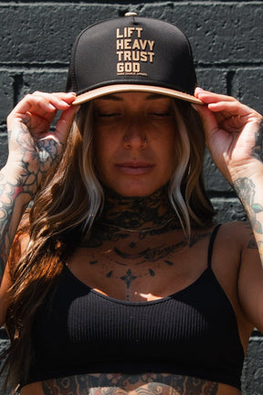 A tattooed woman with long hair adjusts the Lift Heavy. Trust God. Hydro - Black Snapback Hat by Tango Charlie Apparel—an iconic, sweat-resistant Christian gym cap—while standing against a dark brick wall in a black ribbed top.