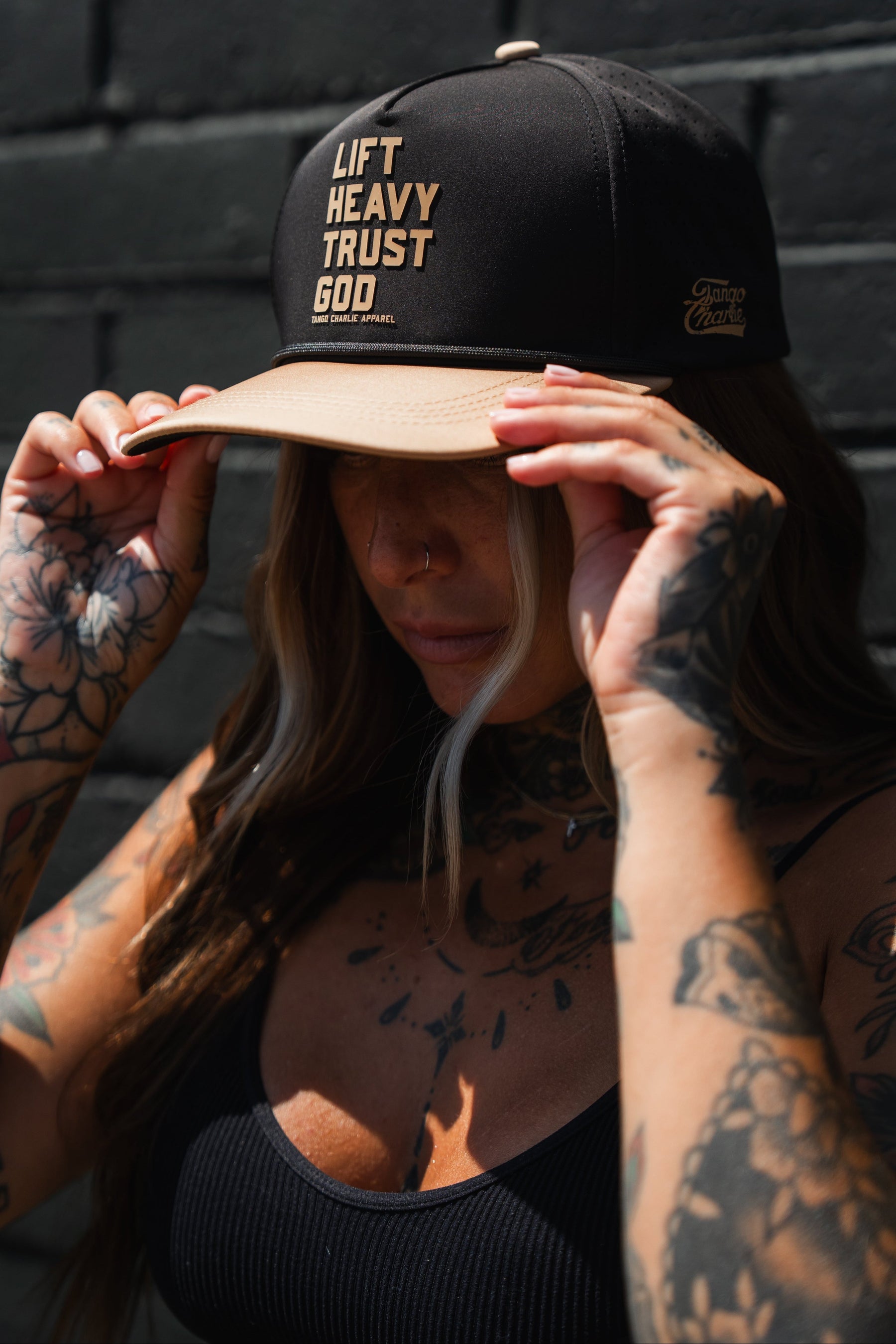 A person with tattoos adjusts a Tango Charlie Apparel Lift Heavy. Trust God. Hydro - Black Snapback Hat, a stylish Christian gym hat featuring sweat-resistant design, while standing against a dark brick wall.