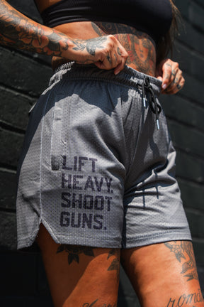 A person wearing Tango Charlie Apparel's Men's Mesh Lifestyle Shorts (5.5" - LHSG OD Green) and a black crop top featuring the text "LIFT. HEAVY. SHOOT. GUNS." stands against a dark brick wall, displaying tattoos on their arms and legs.