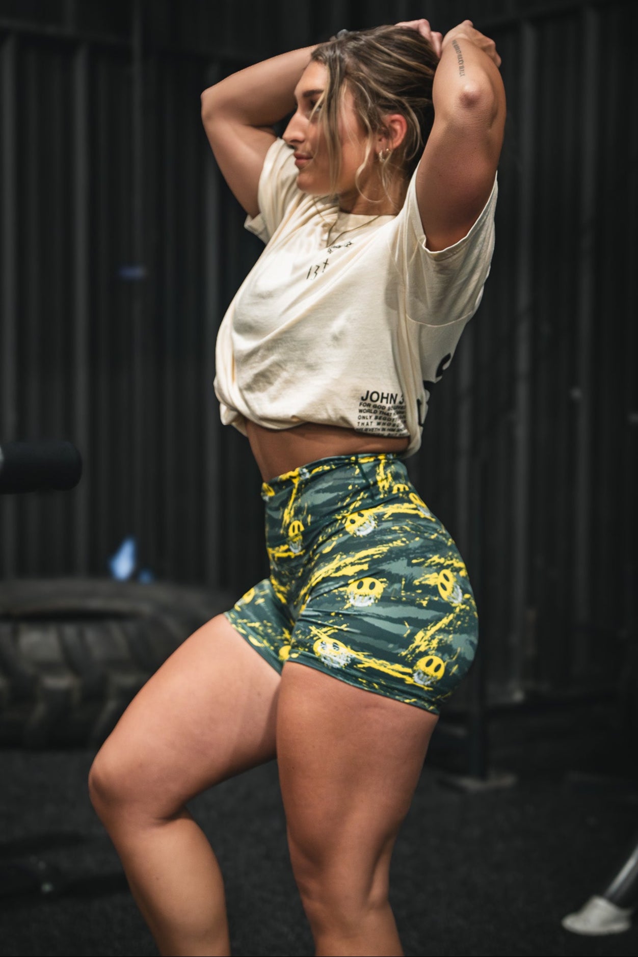 A woman poses in a gym, hands behind her head, wearing a light cropped tee and Tango Charlie Apparel’s Women's High-Waisted Seamless Front Compression Training Shorts | Savage, featuring yellow smiley faces, with gym equipment and a large tire behind her.