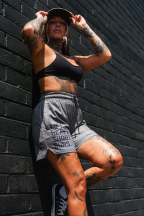 A tattooed person in a black sports bra, Tango Charlie Apparel Men's Mesh Lifestyle Shorts 5.5" - LHSG OD Green, and sneakers poses against a black brick wall, adjusting their cap in sunlight.