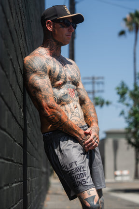A tattooed man wearing sunglasses, a cap, and Tango Charlie Apparel's Men's Mesh Lifestyle Shorts (5.5" - LHSG OD Green) stands shirtless against a black brick wall outdoors on a sunny day.
