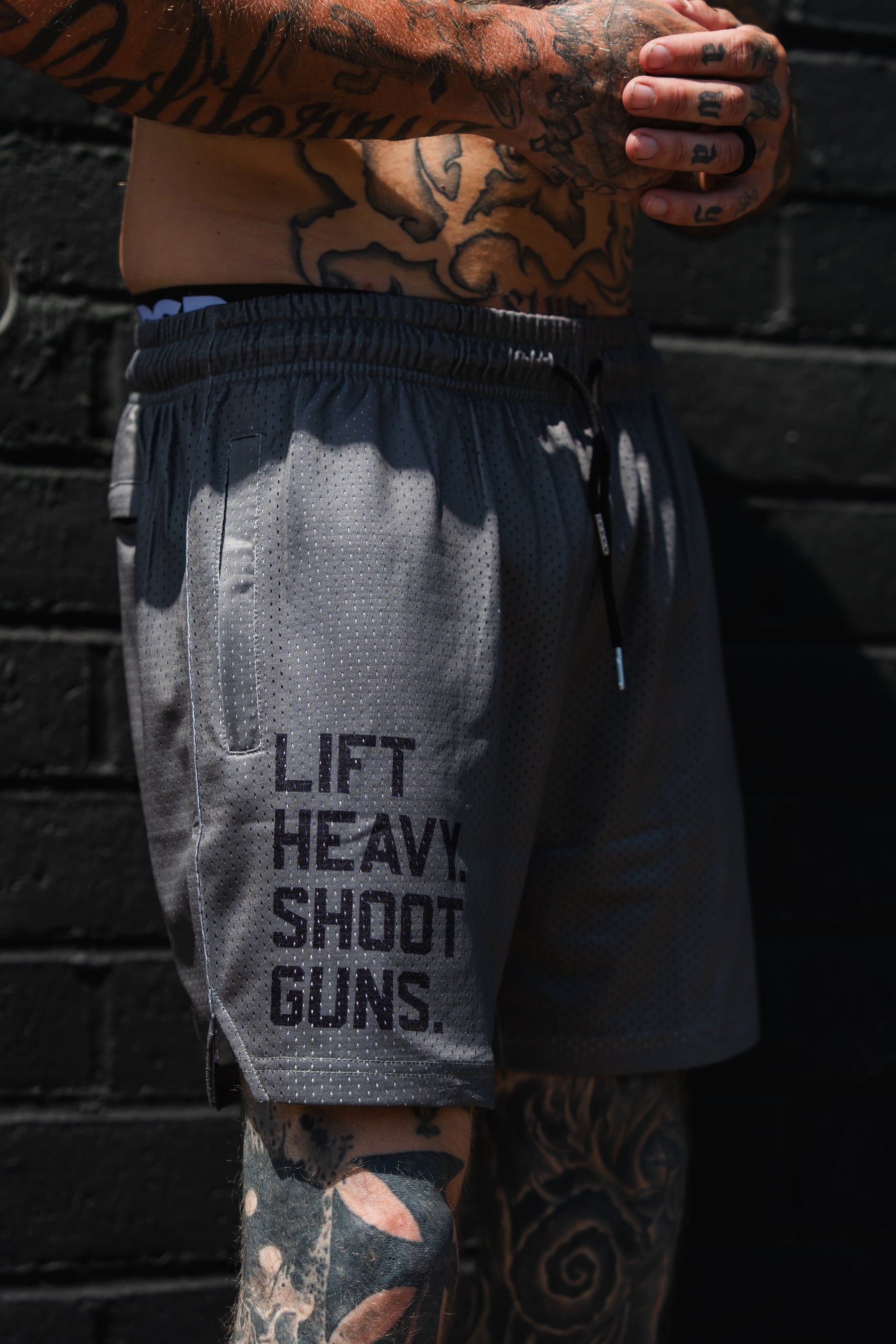 A tattooed individual stands against a black brick wall, sporting the Tango Charlie Apparel Men's Mesh Lifestyle Shorts | 5.5"- LHSG OD Green, featuring “LIFT HEAVY. SHOOT GUNS.” printed on the left leg—a perfect blend of comfort and attitude for an active lifestyle.