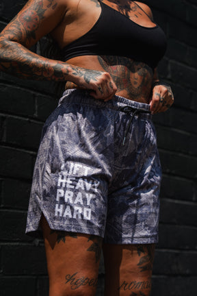 A person with tattoos stands against a dark brick wall, wearing a black sports bra and the Tango Charlie Apparel Men's Mesh Lifestyle Shorts | 5.5"- Lift Heavy Pray Hard Deadwood.