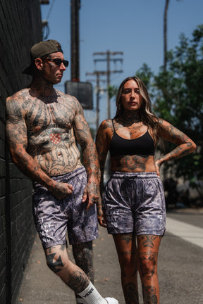 Two heavily tattooed people stand outdoors in athletic wear, including the Men's Mesh Lifestyle Shorts | 5.5" - Lift Heavy Pray Hard Deadwood by Tango Charlie Apparel, posing confidently by a black wall on a sunny day.