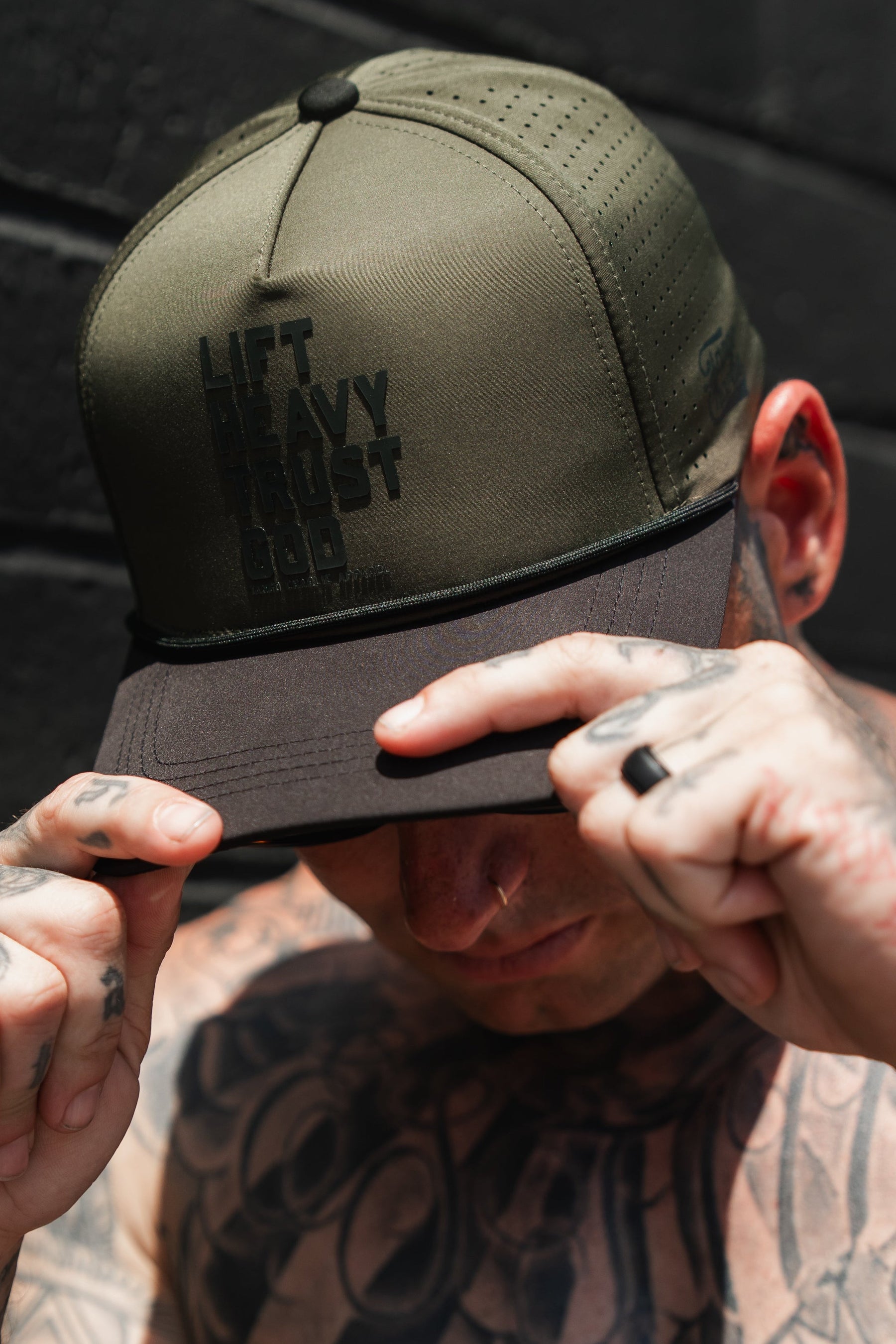 A tattooed man stands against a dark background, gripping the brim of a Tango Charlie Apparel Lift Heavy. Trust God. Hydro - OD Green Snapback Hat with both hands.