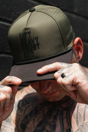 A tattooed man stands against a dark background, gripping the brim of a Tango Charlie Apparel Lift Heavy. Trust God. Hydro - OD Green Snapback Hat with both hands.