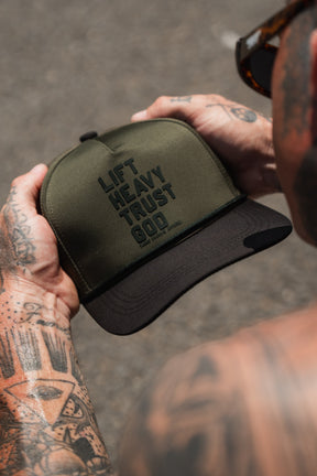 A tattooed individual holds the Tango Charlie Apparel Lift Heavy. Trust God. Hydro - OD Green Snapback Hat, featuring bold “Lift Heavy Trust God” lettering on the front. This water-resistant hat is ideal for intense workouts, keeping you comfortable while expressing your faith.