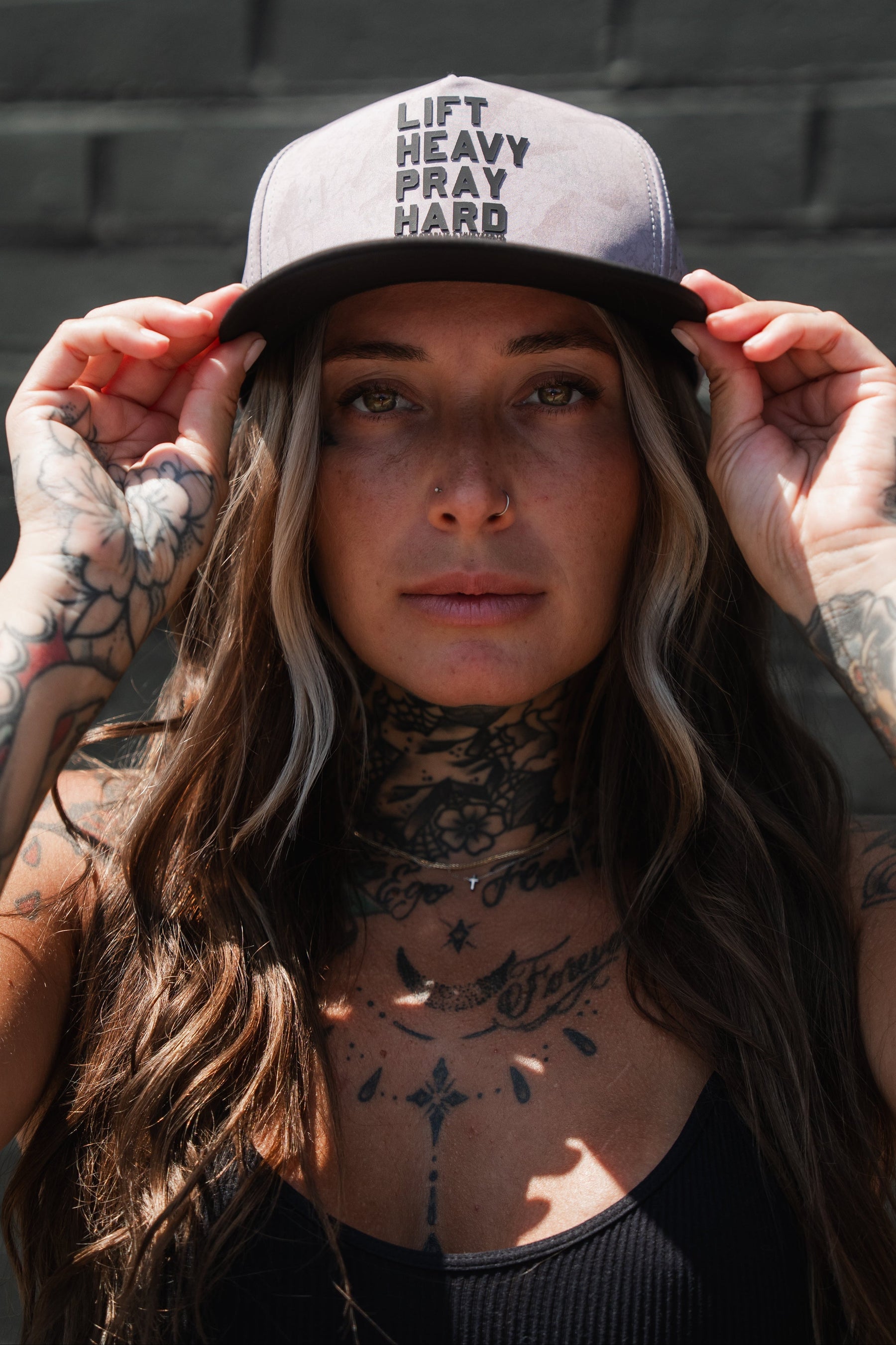 A woman with long hair and tattoos adjusts a Tango Charlie Apparel Lift Heavy. Pray Hard. Deadwood Camo Hydro Snapback Hat while standing against a dark brick wall. This water-resistant Hydro hat offers the perfect blend of style and durability, making it ideal for tough workouts.