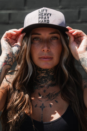 A woman with long hair and tattoos adjusts a Tango Charlie Apparel Lift Heavy. Pray Hard. Deadwood Camo Hydro Snapback Hat while standing against a dark brick wall. This water-resistant Hydro hat offers the perfect blend of style and durability, making it ideal for tough workouts.