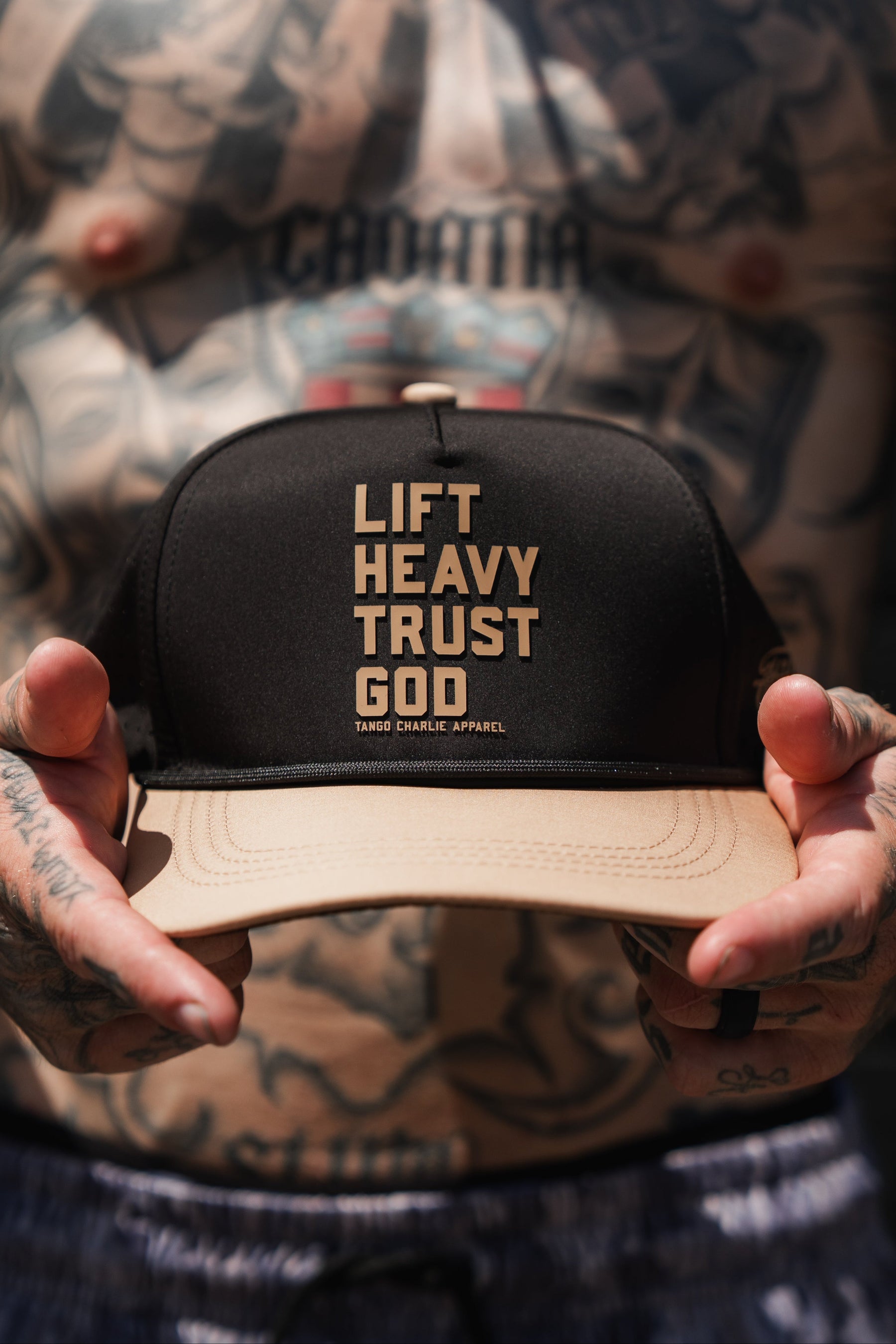 A tattooed person holds the Lift Heavy. Trust God. Hydro - Black Snapback Hat by Tango Charlie Apparel, a Christian gym hat that features LIFT HEAVY TRUST GOD in bold lettering on the front panel.