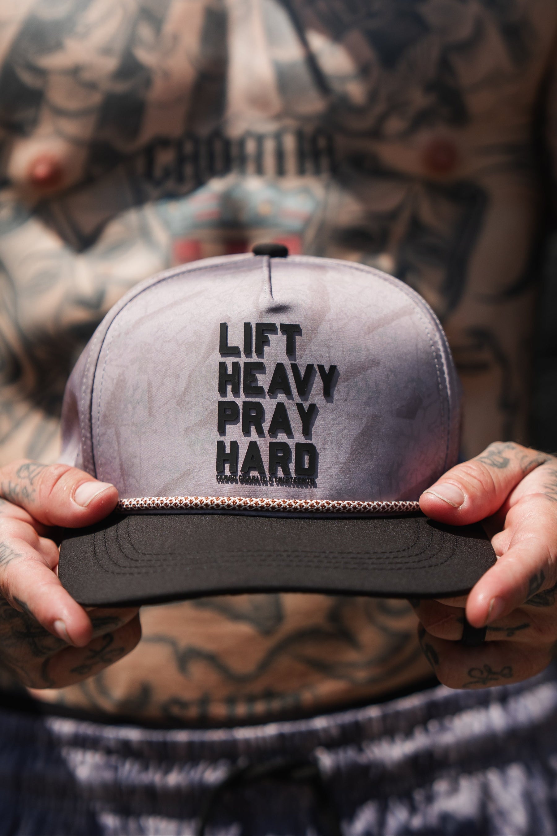 A tattooed individual holds the Lift Heavy. Pray Hard. Deadwood Camo Hydro – Snapback Hat by Tango Charlie Apparel, featuring bold black “LIFT HEAVY PRAY HARD” lettering on the front. This water-resistant Hydro hat is built to handle your toughest workouts and daily adventures.