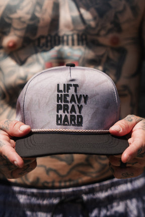 A tattooed individual holds the Lift Heavy. Pray Hard. Deadwood Camo Hydro – Snapback Hat by Tango Charlie Apparel, featuring bold black “LIFT HEAVY PRAY HARD” lettering on the front. This water-resistant Hydro hat is built to handle your toughest workouts and daily adventures.