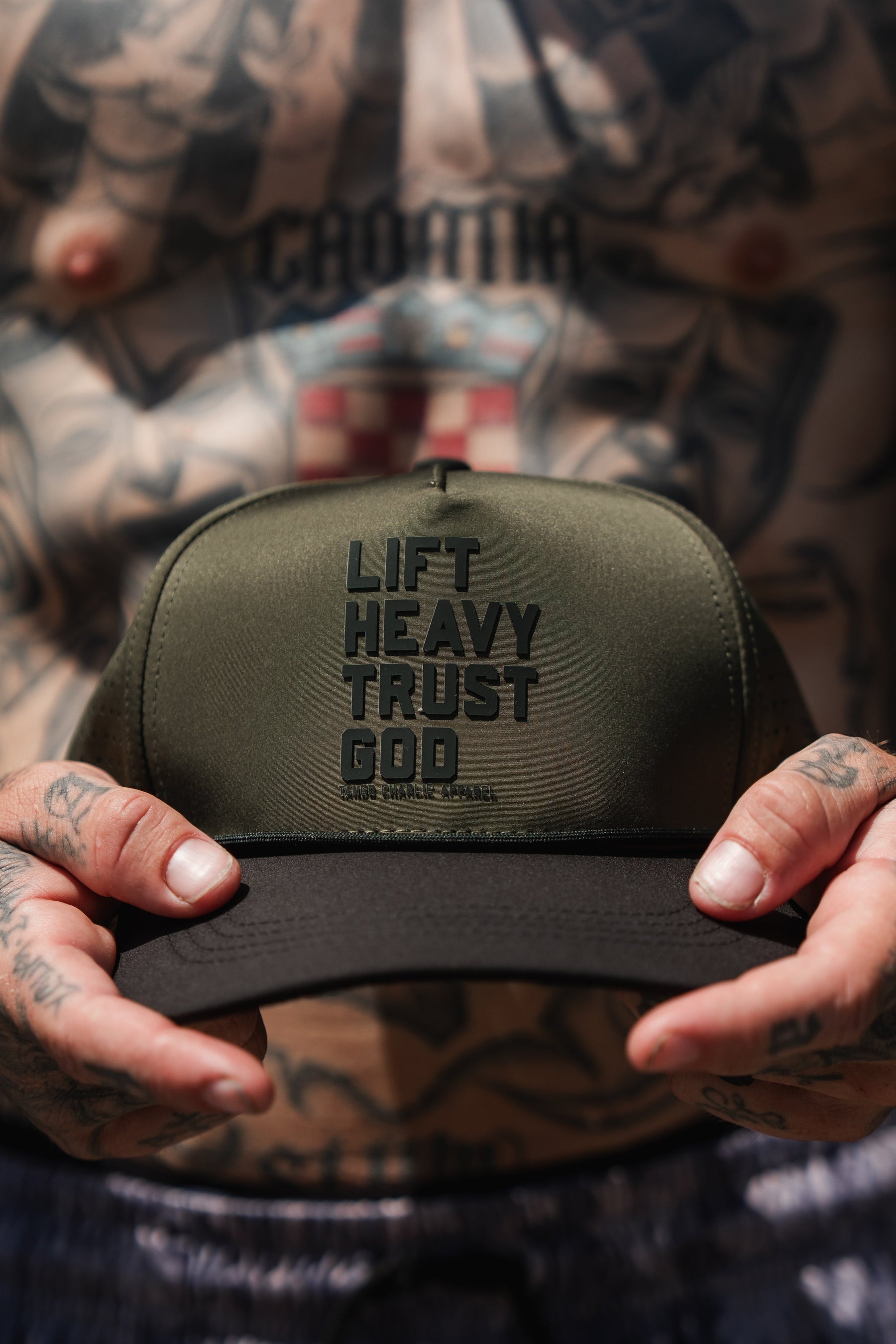 A tattooed individual holds the Tango Charlie Apparel Lift Heavy. Trust God. Hydro - OD Green Snapback Hat, a Christian gym hat in olive green and black that’s sweat-resistant—perfect for workouts and faith-driven motivation.