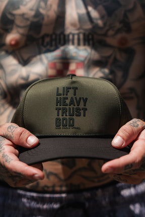 A tattooed individual holds the Tango Charlie Apparel Lift Heavy. Trust God. Hydro - OD Green Snapback Hat, a Christian gym hat in olive green and black that’s sweat-resistant—perfect for workouts and faith-driven motivation.