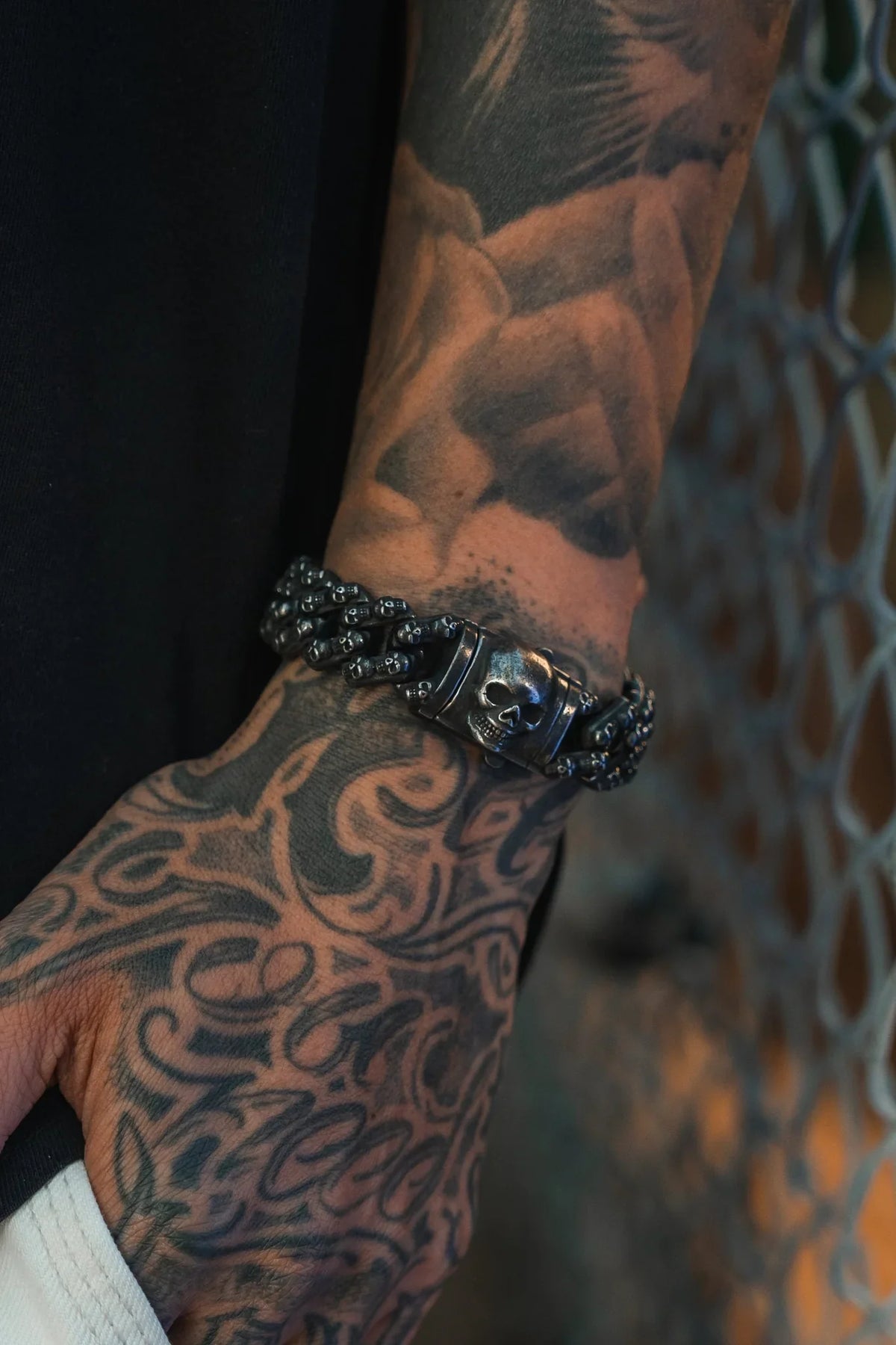 A tattooed hand and forearm showcase the Skulled Bracelet by Relentless Betrayal, its bold dark metal gleaming against a metal fence background.