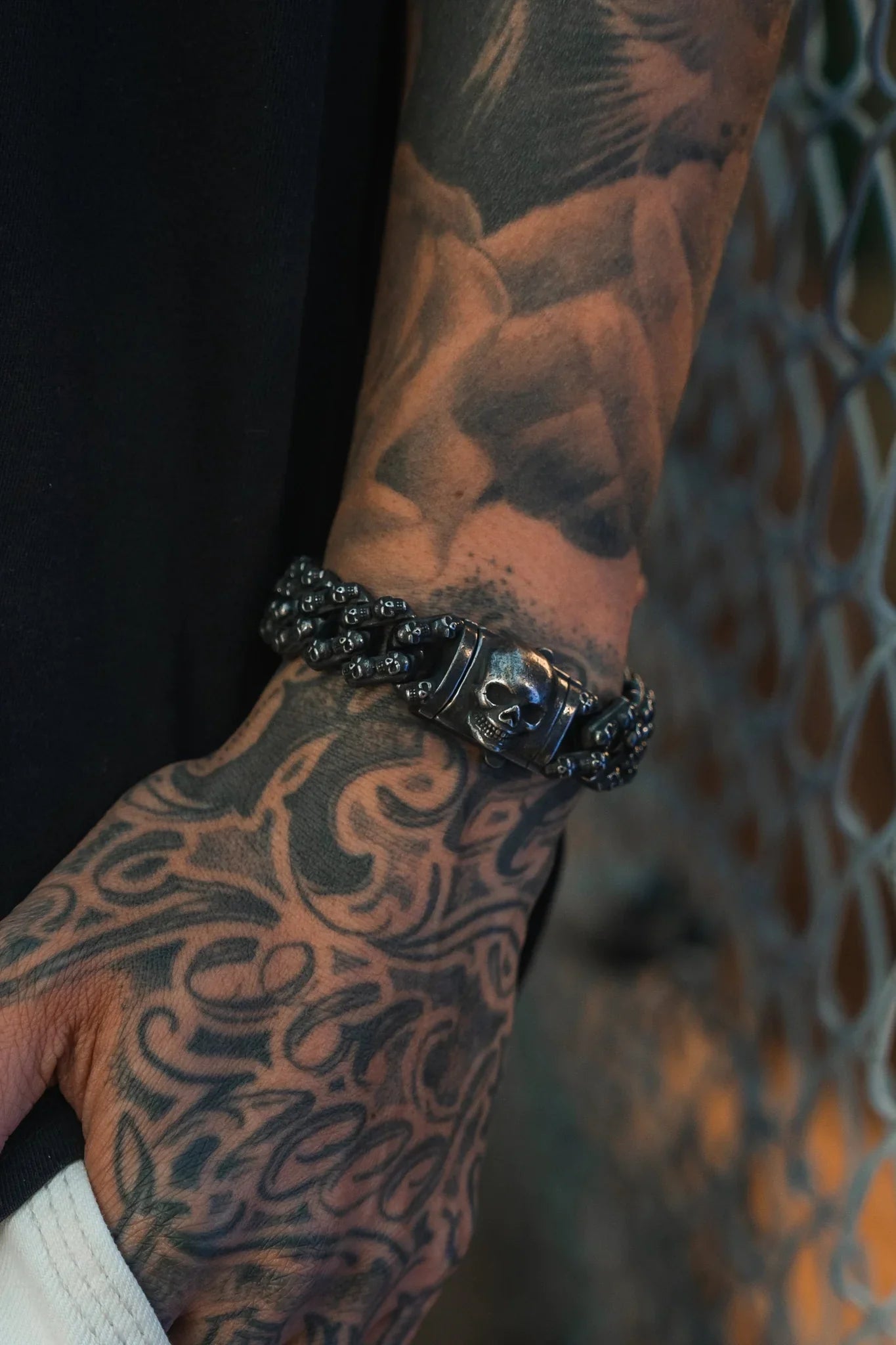 A tattooed hand and forearm showcase the Skulled Bracelet by Relentless Betrayal, its bold dark metal gleaming against a metal fence background.