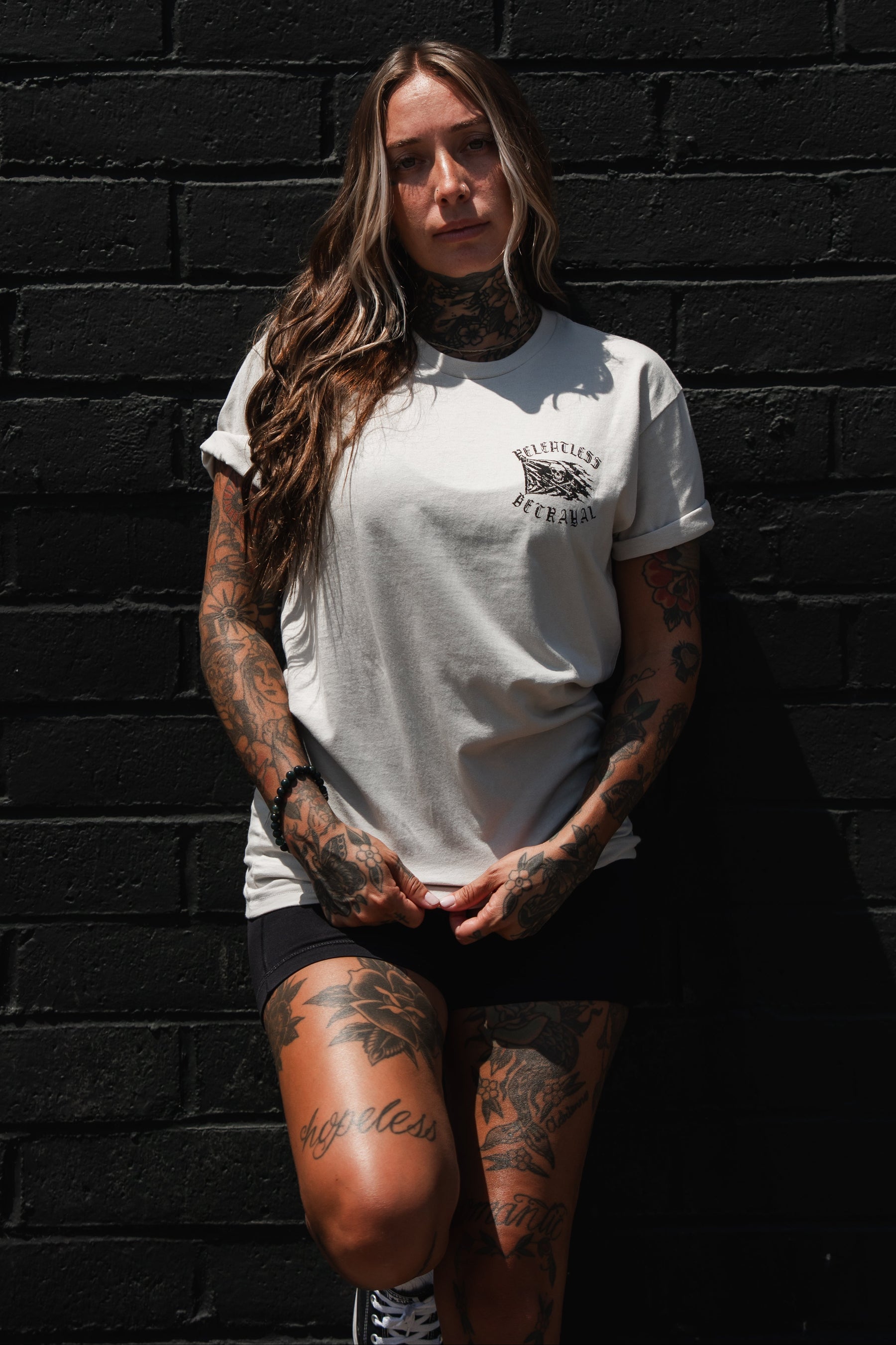 A woman with long hair and tattoos stands against a black brick wall, wearing the Relentless Betrayal “Raise the Jolly Roger Bone” T-Shirt and black shorts—a nod to pirate streetwear—as she looks at the camera with her hands clasped.