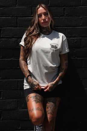 A woman with long hair and tattoos stands against a black brick wall, wearing the Relentless Betrayal “Raise the Jolly Roger Bone” T-Shirt and black shorts—a nod to pirate streetwear—as she looks at the camera with her hands clasped.