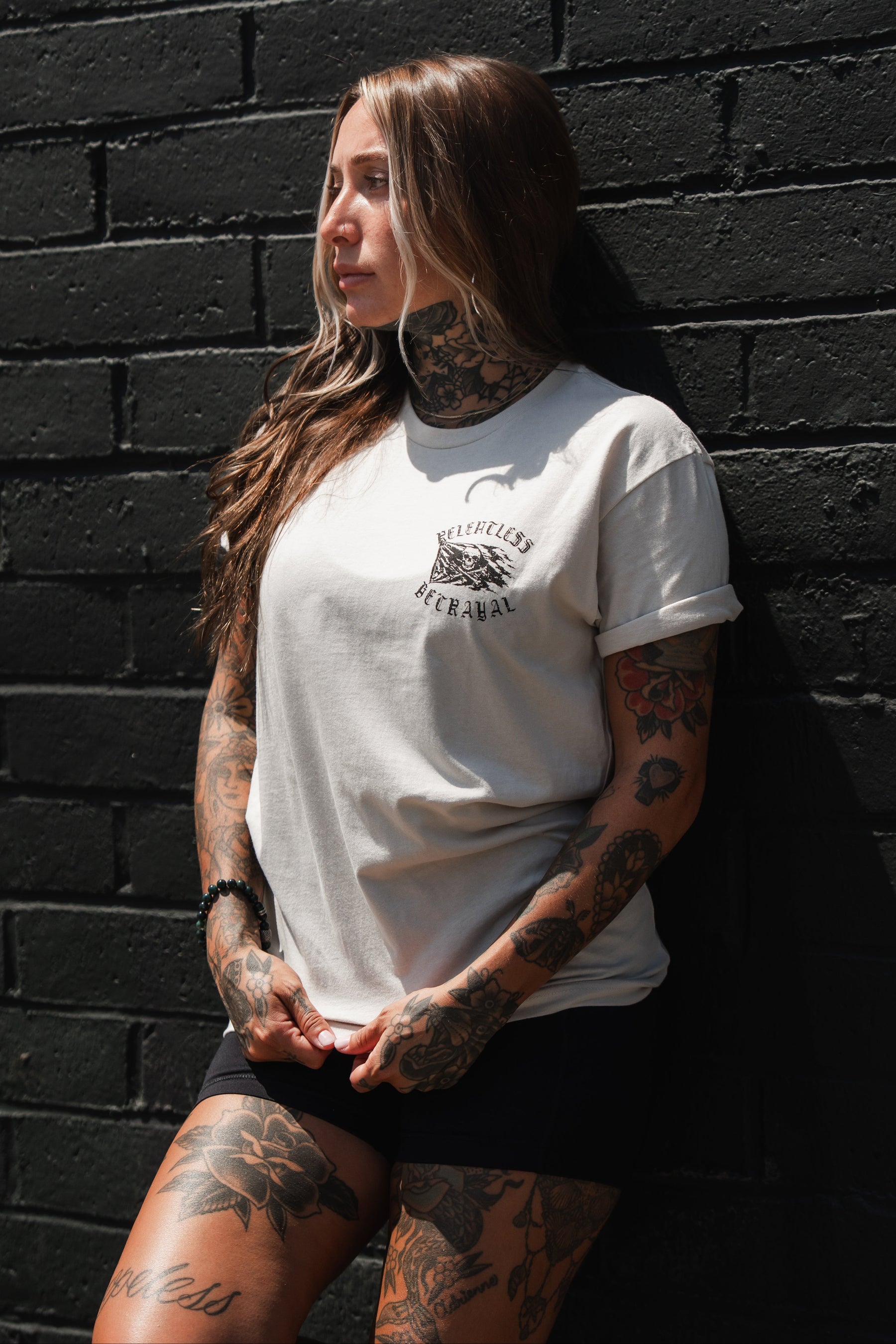 A tattooed woman in the Relentless Betrayal Raise the Jolly Roger Bone T-Shirt and black shorts stands against a black brick wall, glancing to the side, her pirate streetwear style exuding effortless boldness.